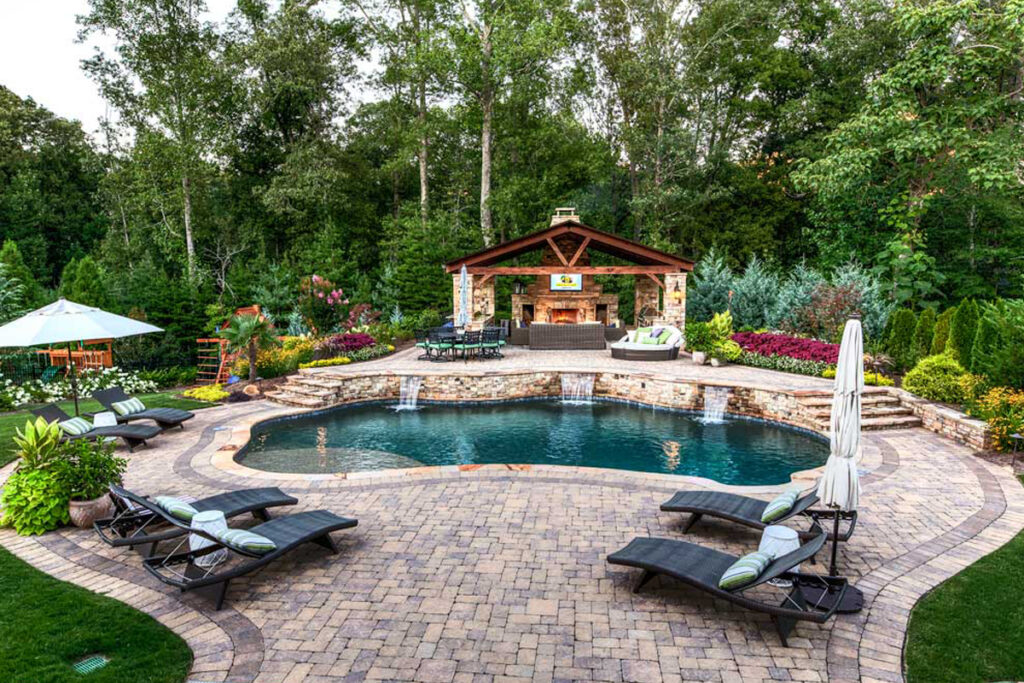 Luxurious residential landscape design with a custom pool, stone patio, outdoor fireplace, and lush greenery. Residential landscaping by DesignX Studios.